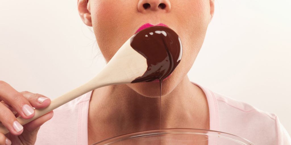 Woman with pink lipstick licking chocolate batter from wooden spoon